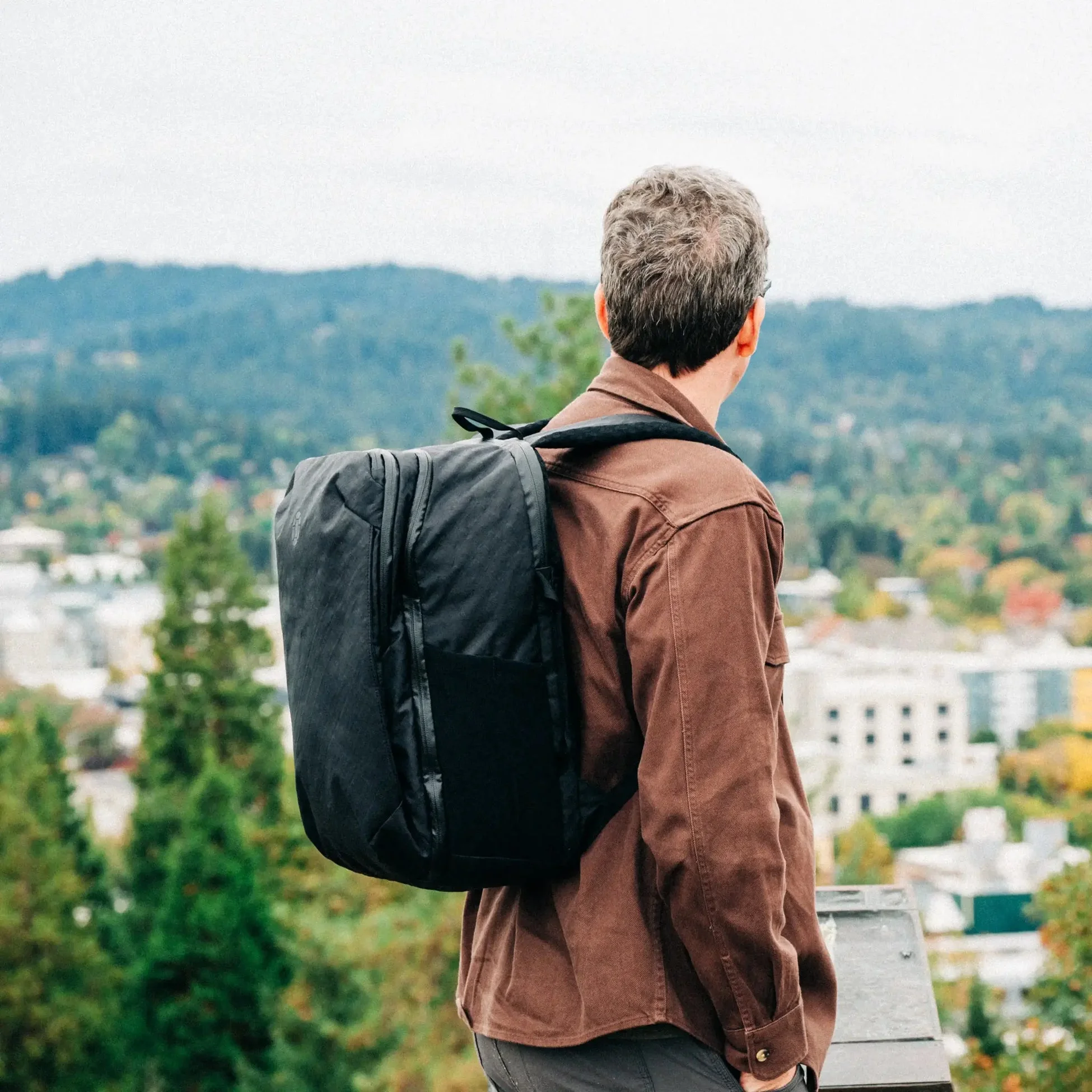 A traveler in a brown jacket looks out over a hillside town with a Tortuga backpack